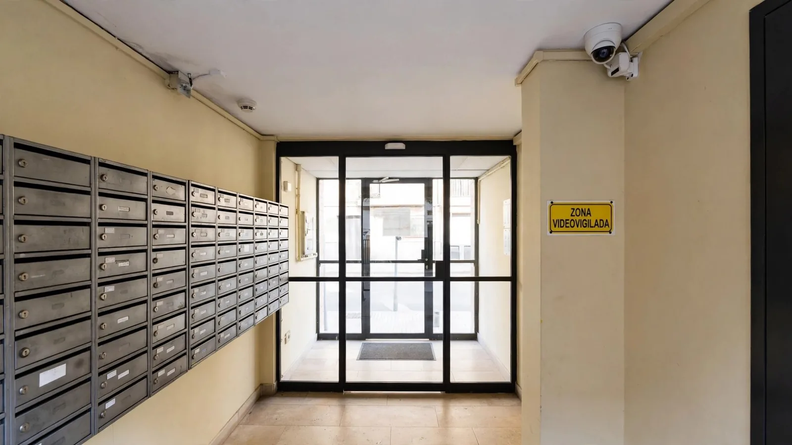 Portal de un edificio de viviendas con buzones metálicos, una cámara de videovigilancia en el techo y un cartel amarillo de zona videovigilada