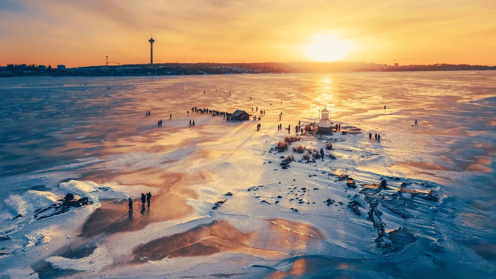 Lago helado al atardecer con gente paseando junto a un pequeño faro y la ciudad de Tampere al fondo
