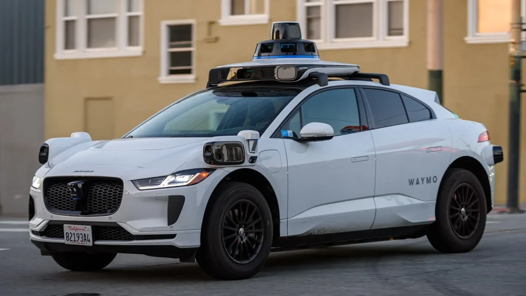 Coche autónomo blanco de Waymo, un Jaguar I-Pace con sensores en el techo y laterales, circulando por una calle